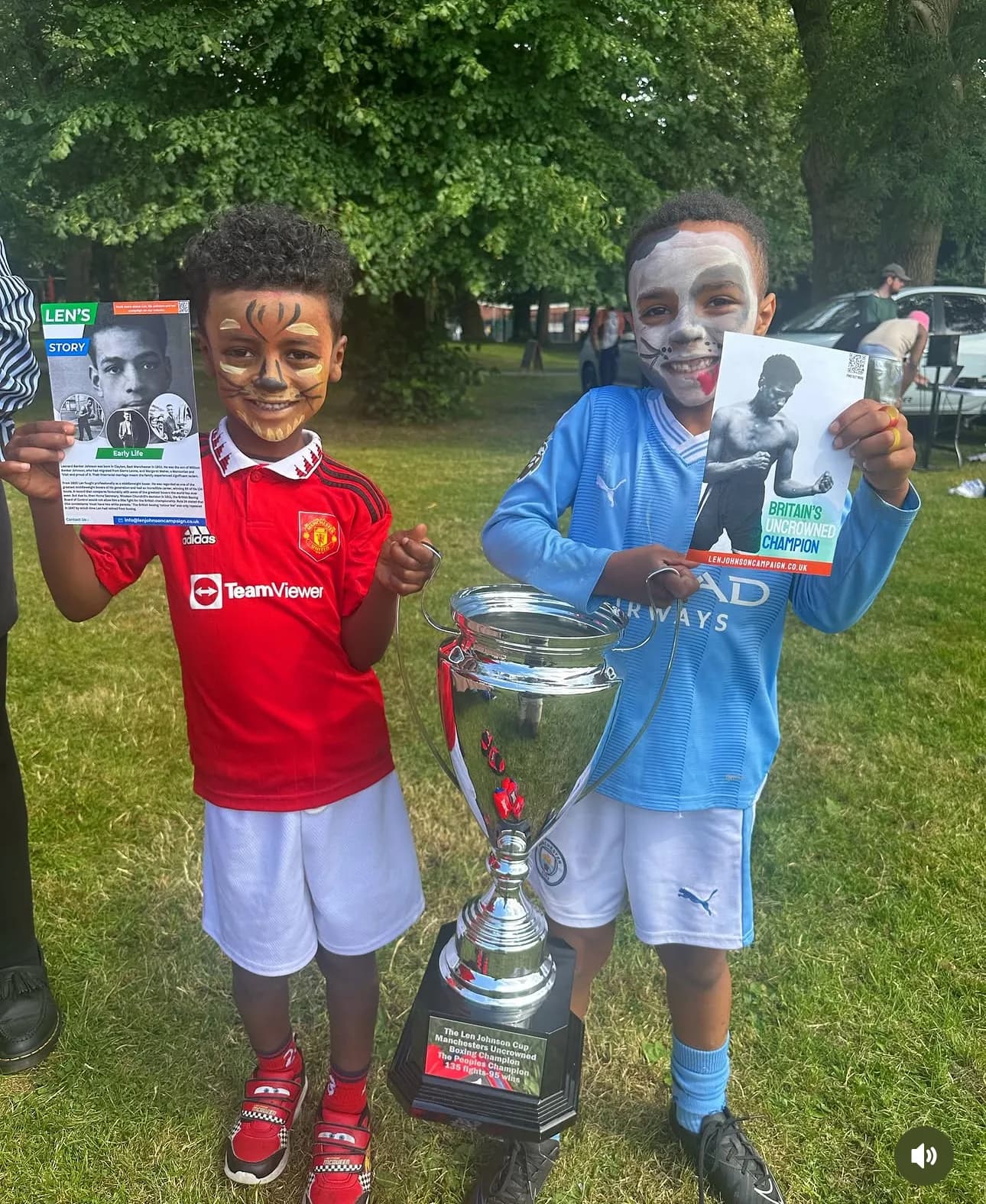 Two children with face paint hold Len Johnson Campaign leaflets beside the Len Johnson Cup trophy in a park