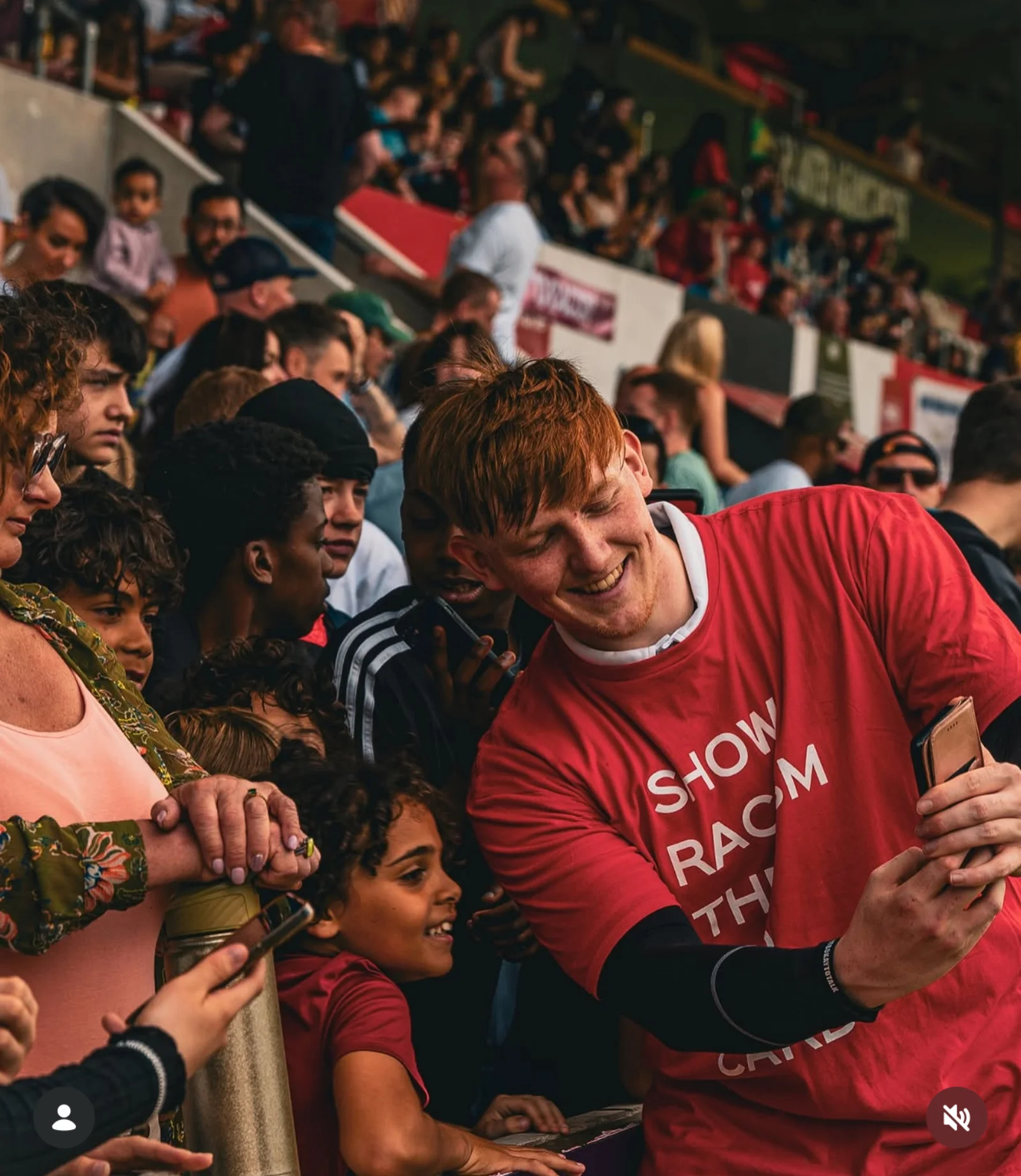 A young fan poses for a selfie wearing a Show Racism the Red Card shirt, surrounded by other spectators in the stand