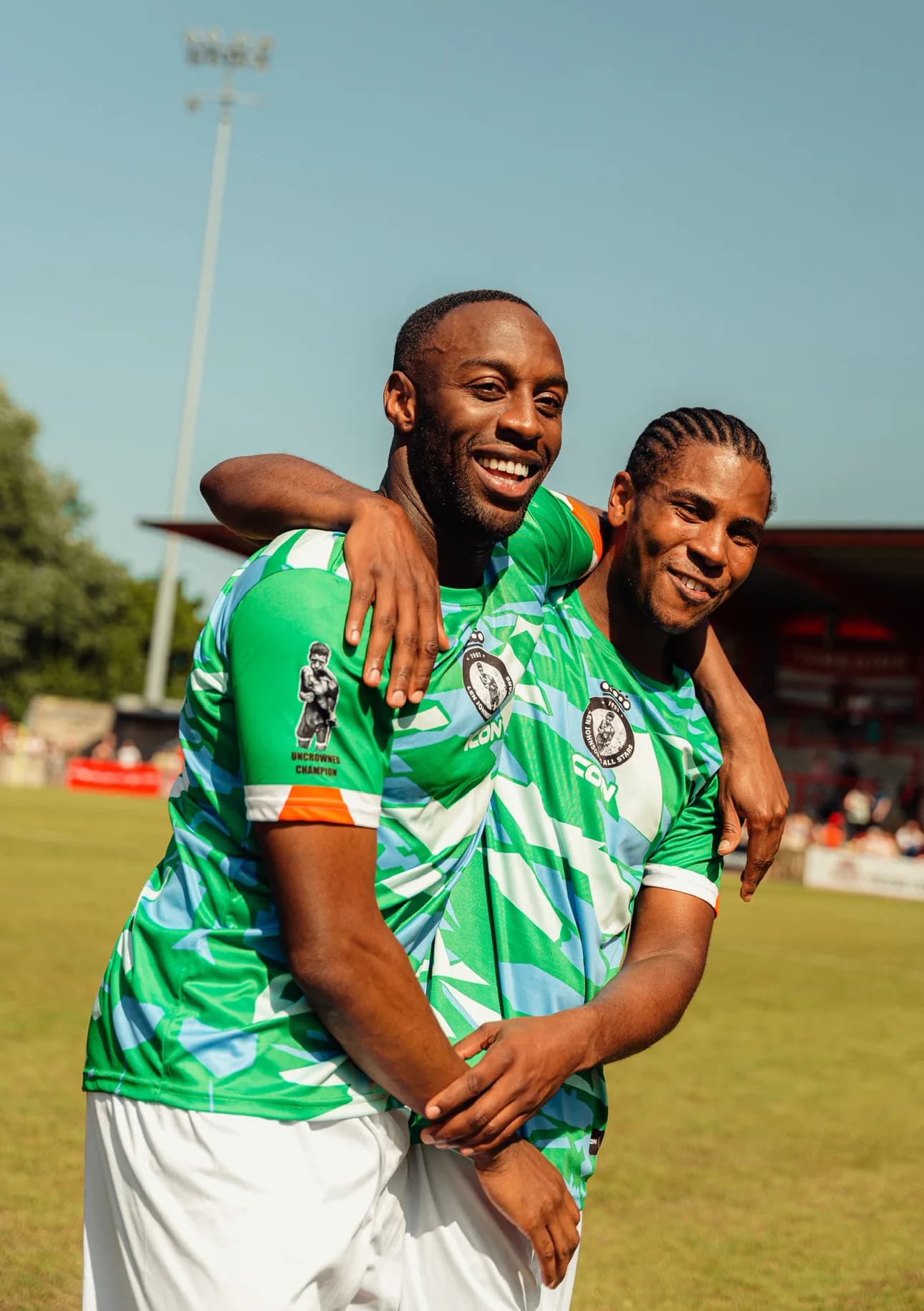 Two players embrace after the match, one wearing a Len Johnson FC All Stars shirt printed with Uncrowned Champion