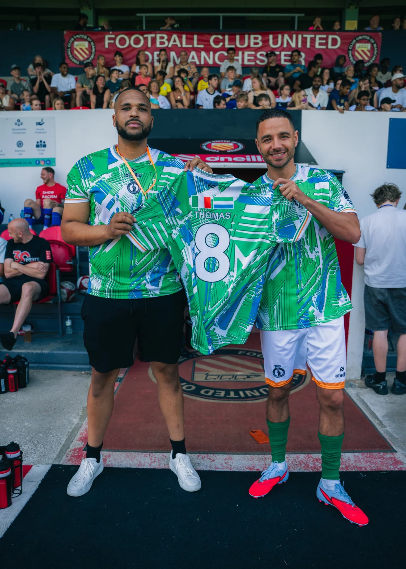 Two players hold up a matchday shirt printed Thomas 8, in front of the Football Club United of Manchester banner