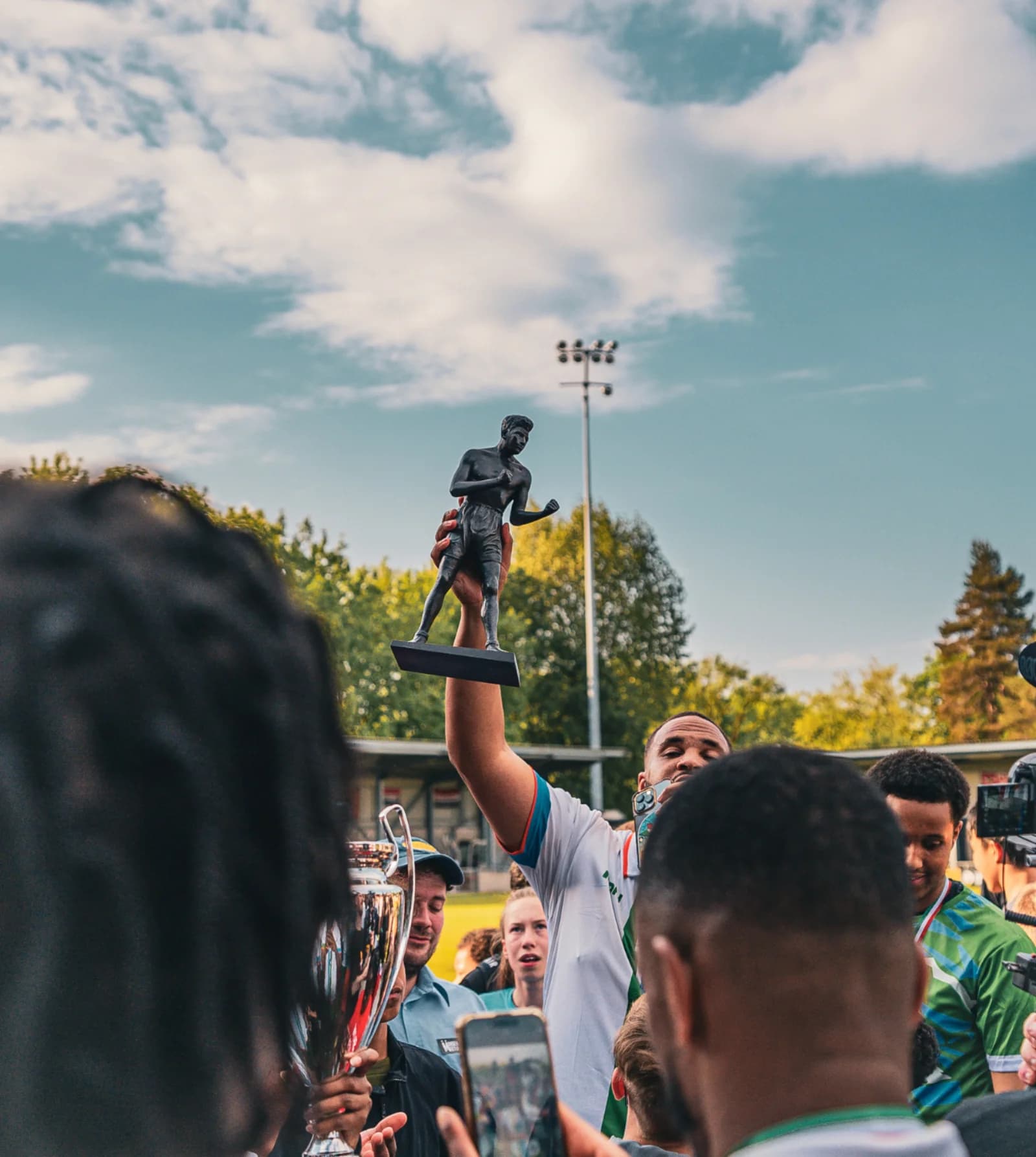 A player holds a small bronze boxer statuette aloft against the sky, pitchside at Broadhurst Park