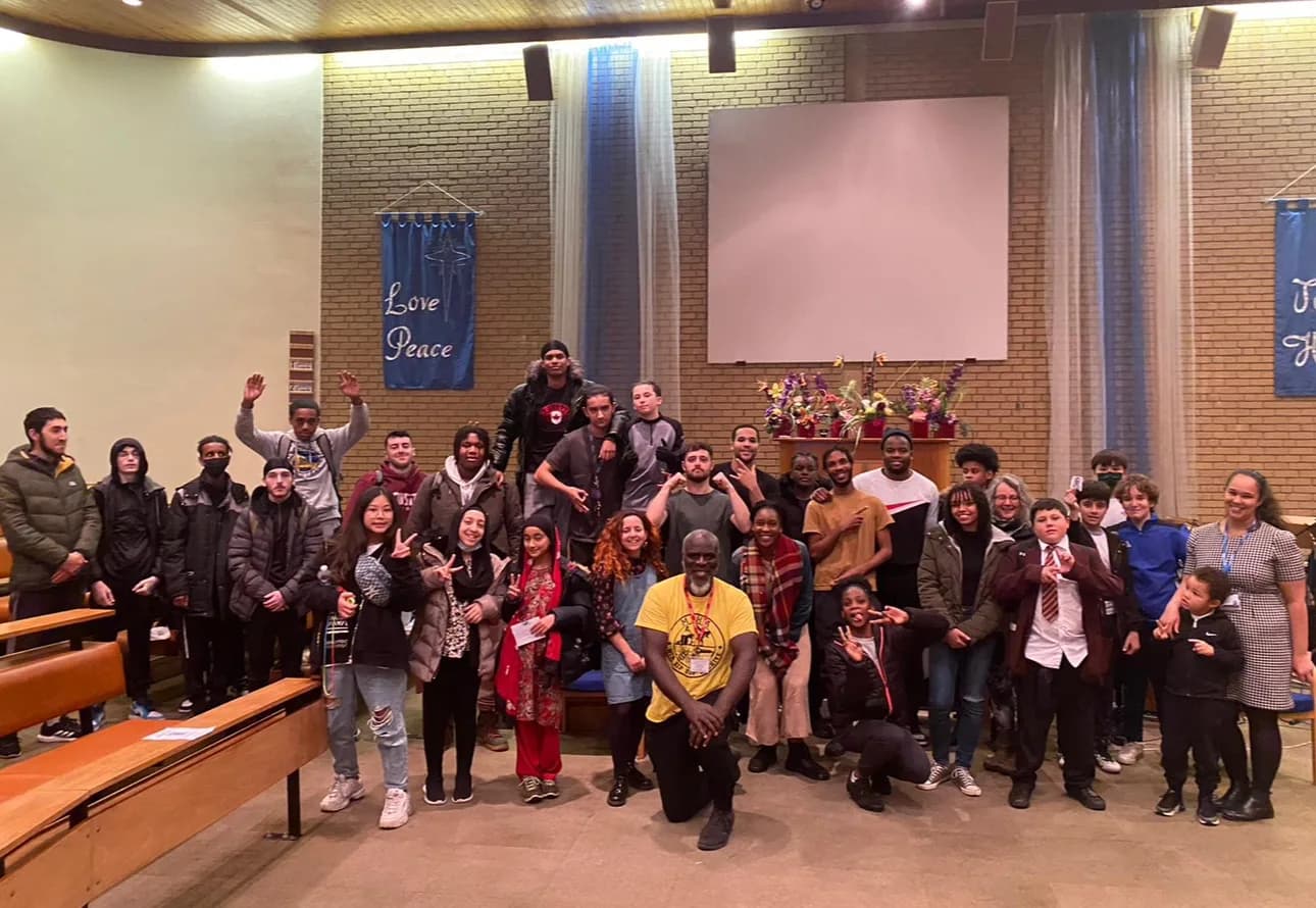 A large group of young people and youth workers pose together in a community hall after a performance, beneath banners reading Love and Peace
