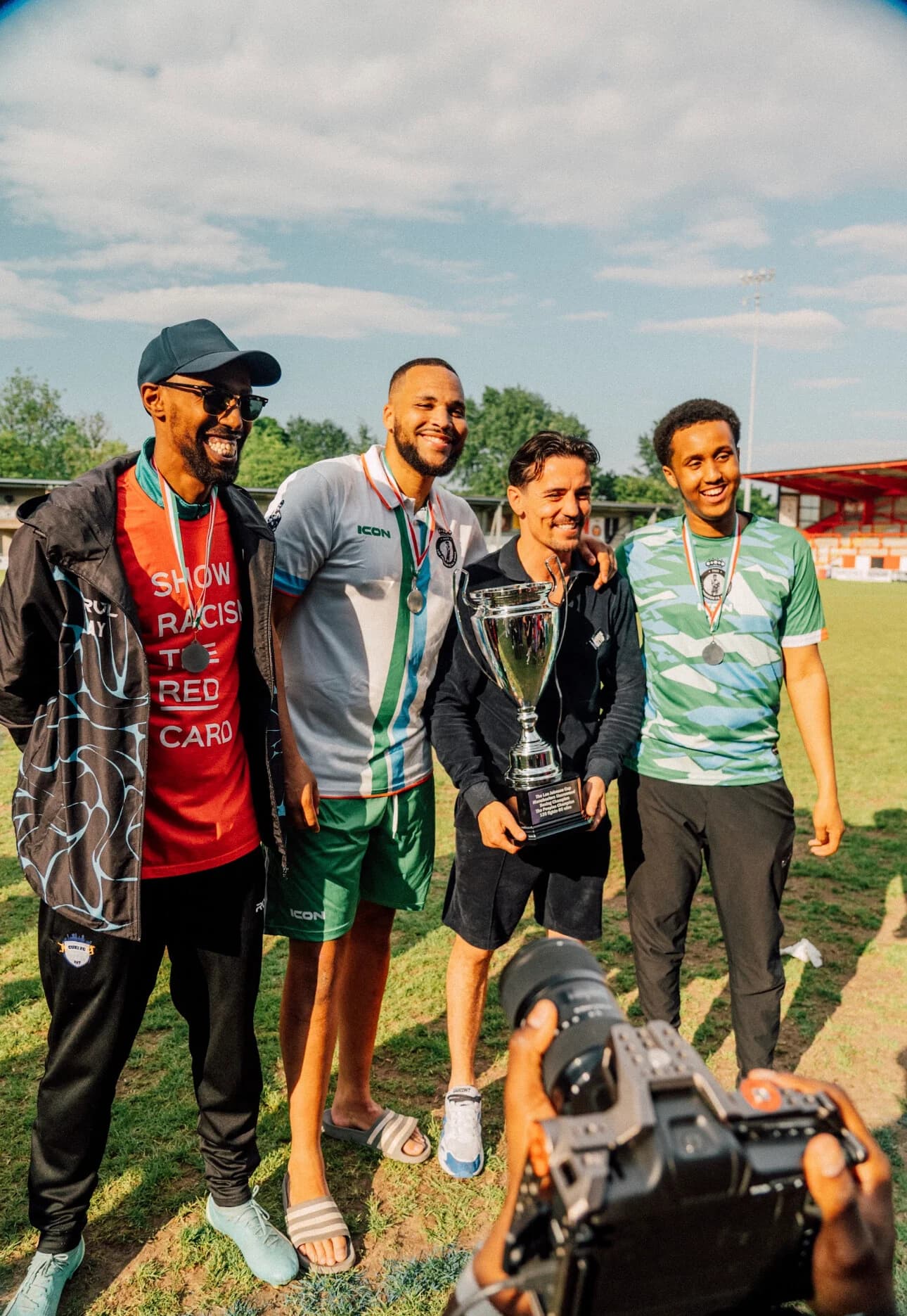 Four men hold a trophy pitchside, one wearing a Show Racism the Red Card shirt