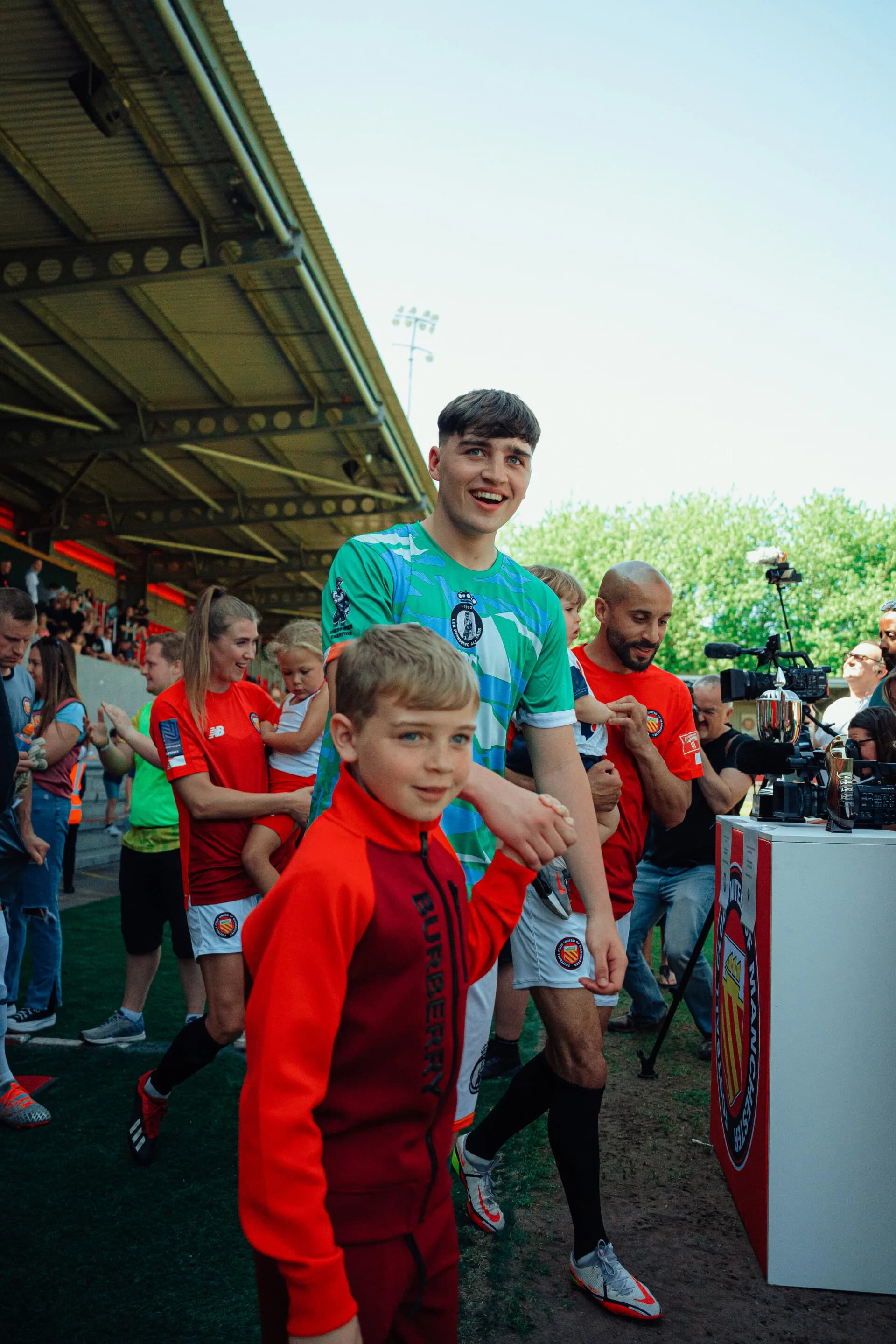 Players from Len Johnson FC All Stars and FC United Legends walk out together at Broadhurst Park, children alongside them
